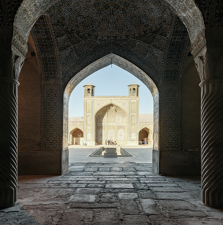 View from within an ornate, arched corridor looking out toward a historic mosque courtyard, featuring intricate tilework, carved columns, and a sunlit entrance with two towers.
