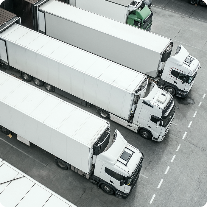 Aerial view of several white semi-trailer trucks parked in a row in a parking lot, with gray pavement and marked parking spaces visible.