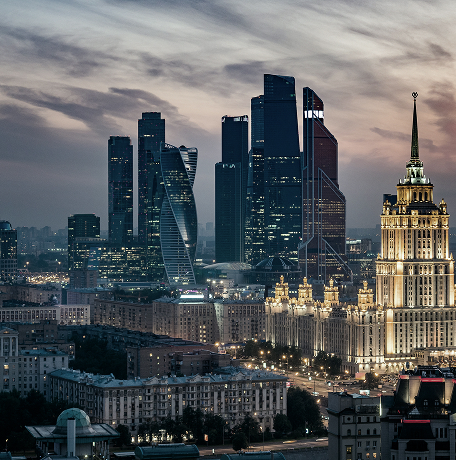 Cityscape at dusk featuring modern skyscrapers and a historic, illuminated building with a spire, under a cloudy sky.