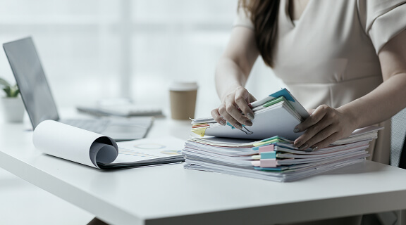 A person sitting at a desk sorting through a stack of papers with colorful tabs, next to a laptop, a coffee cup, and rolled-up documents in a bright office setting.