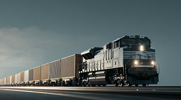A long freight train with stacked cargo containers moves along railroad tracks under a cloudy sky, with the locomotives headlights illuminated.